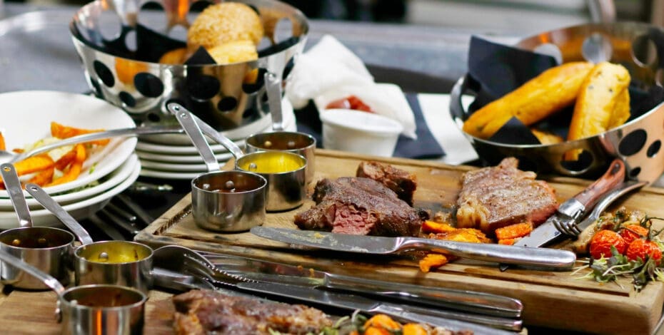 Food waste on wooden boards on a restaurant table. The food waste is mostly half-eaten steak, roasted tomatoes, and roasted carrots.